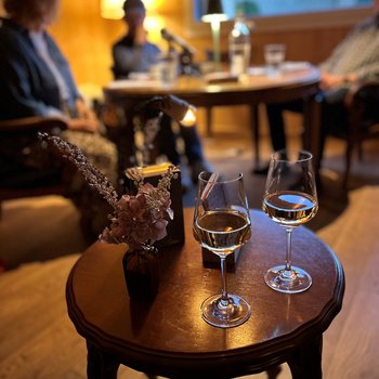 Eins Salontisch mit zwei Weingläser stehen im Vordergrund. Im Hintergrund ist die Gesprächsrunde an einem grossen runden Tisch verschwommen wahrnehmbar