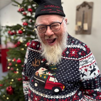 Lachender Mann mit Weihnachtspulli und Mütze vor Weihnachtsbaum
